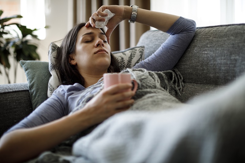 Woman sick on couch holding a mug and a tissue with her hand on her forehead.