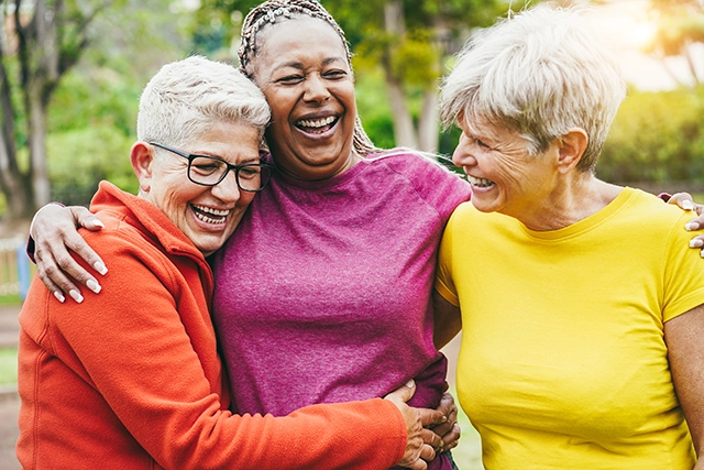 Three aged women embrace in the middle of an outdoor park. They're each wearing different-colored solid sweaters.