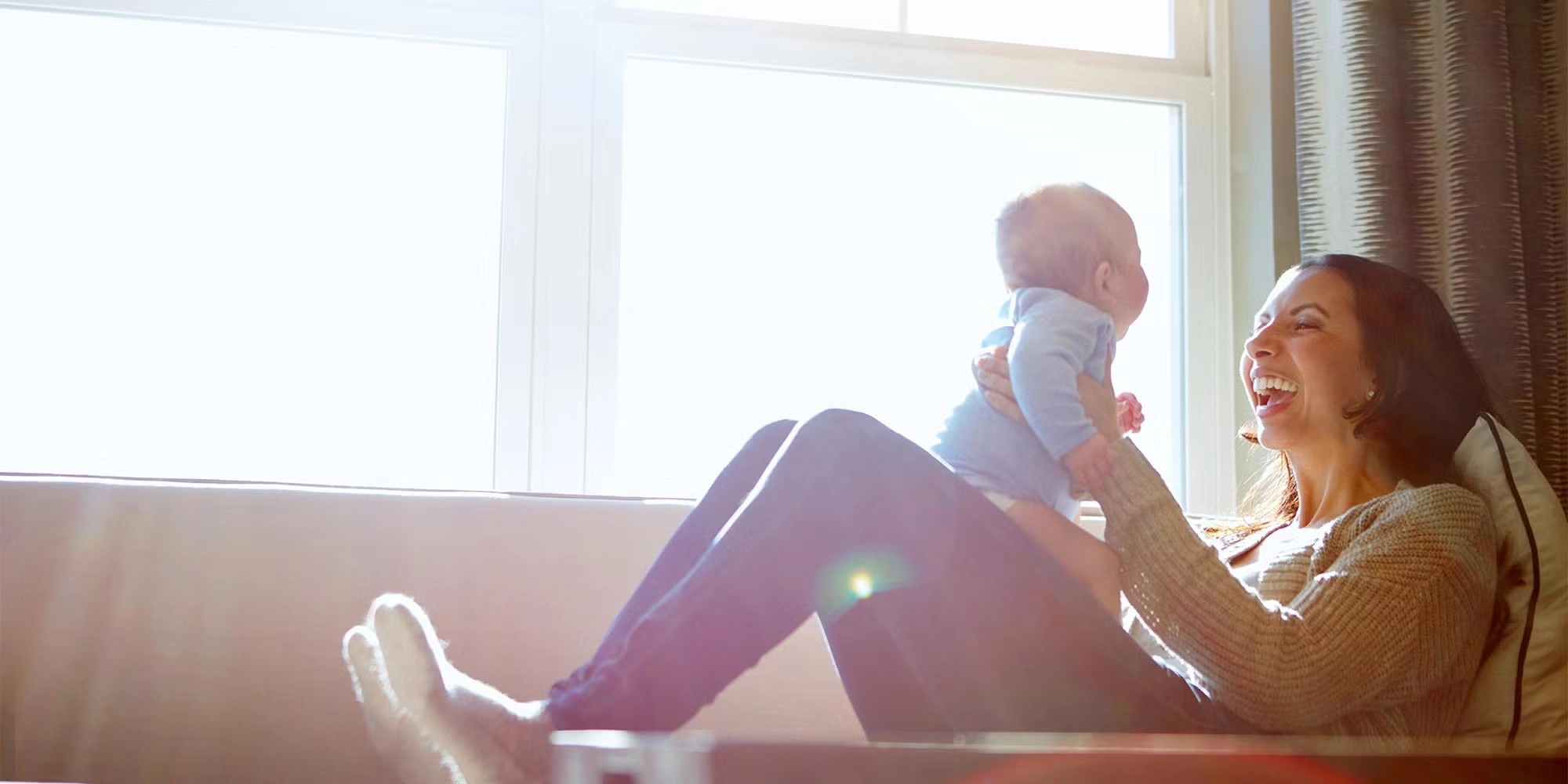 A young woman holds her newborn on her knees. They're playing together. Both are on a tan couch which is backlit by sunshine coming through a window.