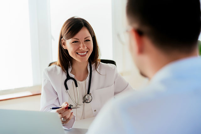 A man sits at a desk and listens to his doctor. She sits across from him smiling, wearing a white coat and stethoscope.
