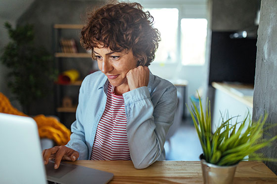 A woman gazes at her computer while scheduling a doctor's appointment. She's inside surrounded by potted plants and wood features.