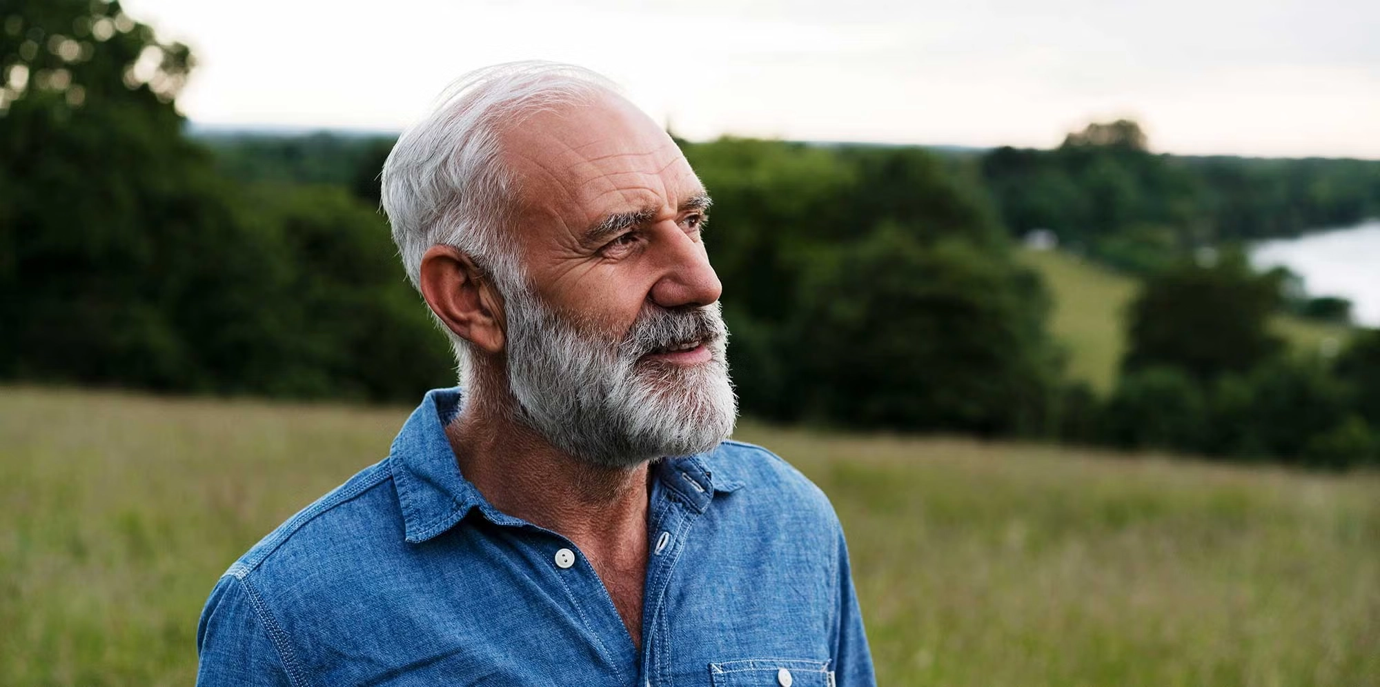 An elder man in a long sleeve blue shirt stands on a grassy hill, looking outward