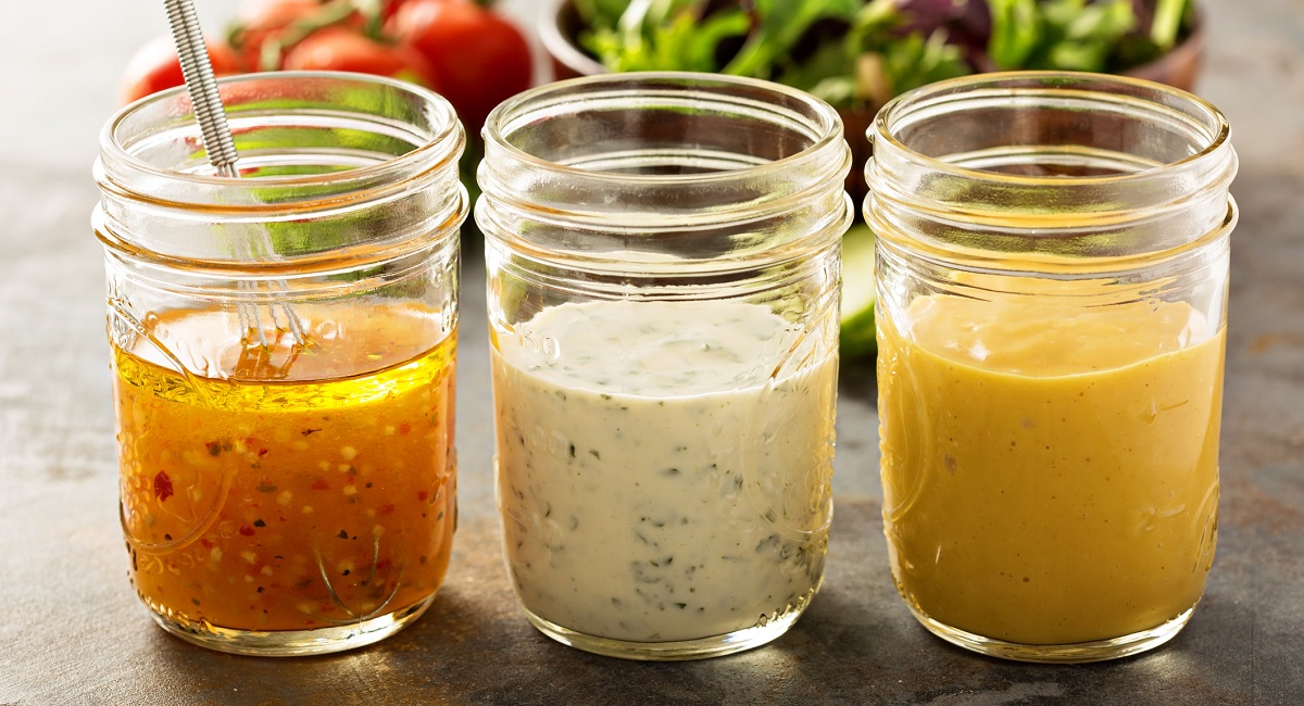 Three small glass jars each filled with a different salad dressing on a granite countertop with tomatoes and salad behind.