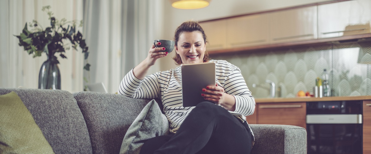 Woman sitting on couch reading a tablet and holding a mug.