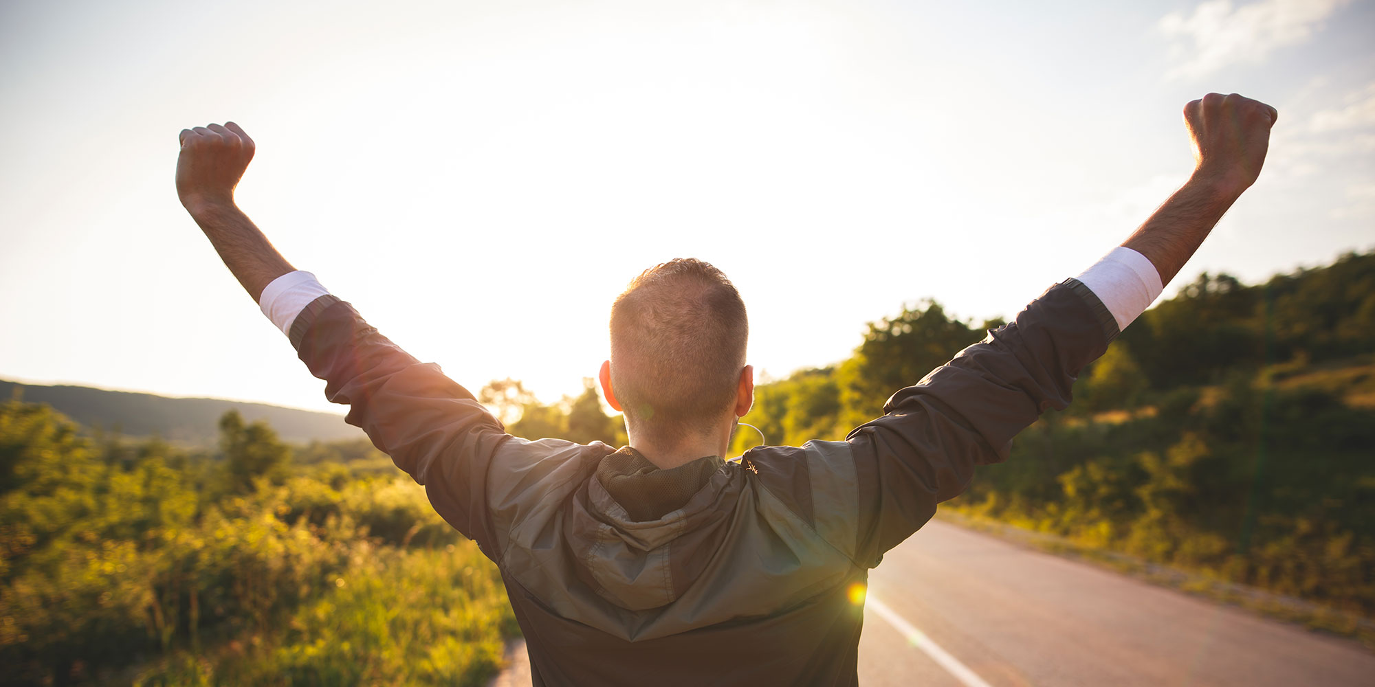 A man lifts his arms in celebration at the end of his run. He's in the middle of a street and the sun is beginning to set.
