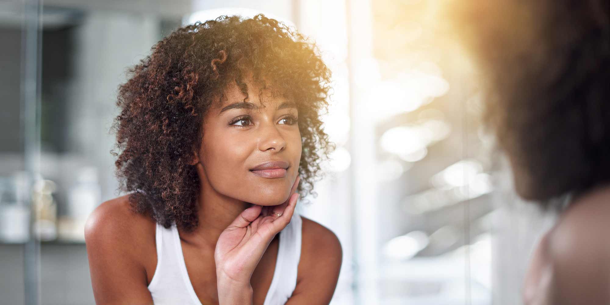A woman glowing in sunlight looks into the mirror with her hand resting on her chin