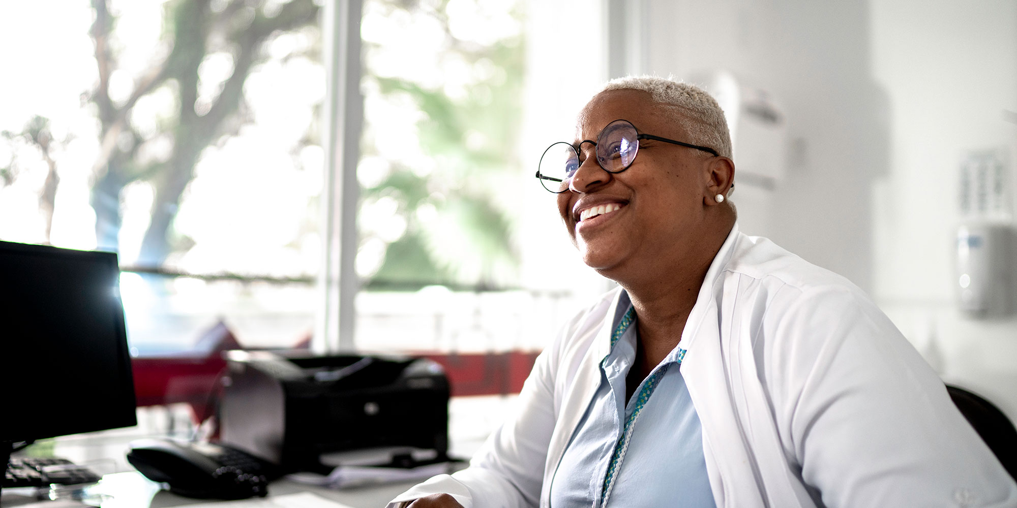 A doctor sits at her desk and smiles as she works on paperwork