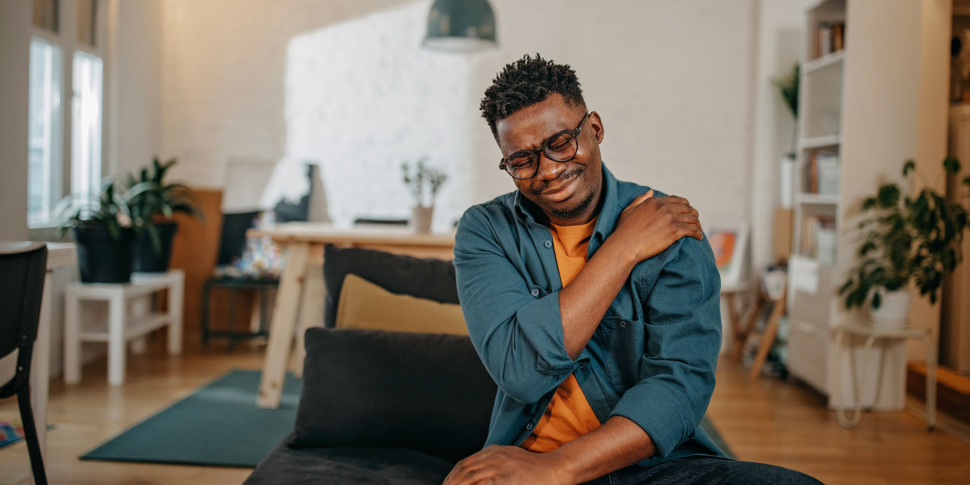 A man winces in pain and holds onto his shoulder. He's sitting in a well-decorated studio apartment.