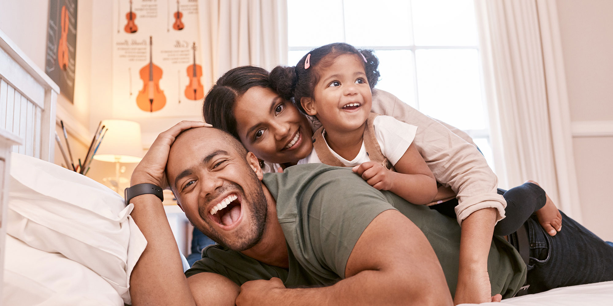 A young father lays on his stomach on a bed, with his happy wife and daughter laying on top of his back