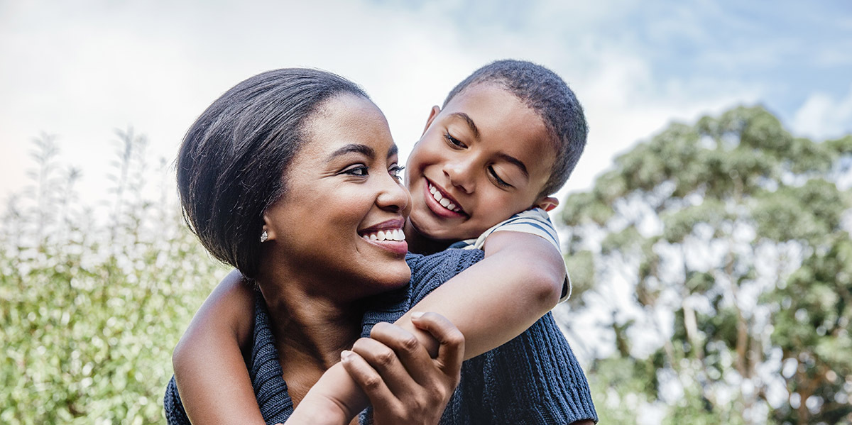 A mother carries her son on her back, smiling together outside in a park