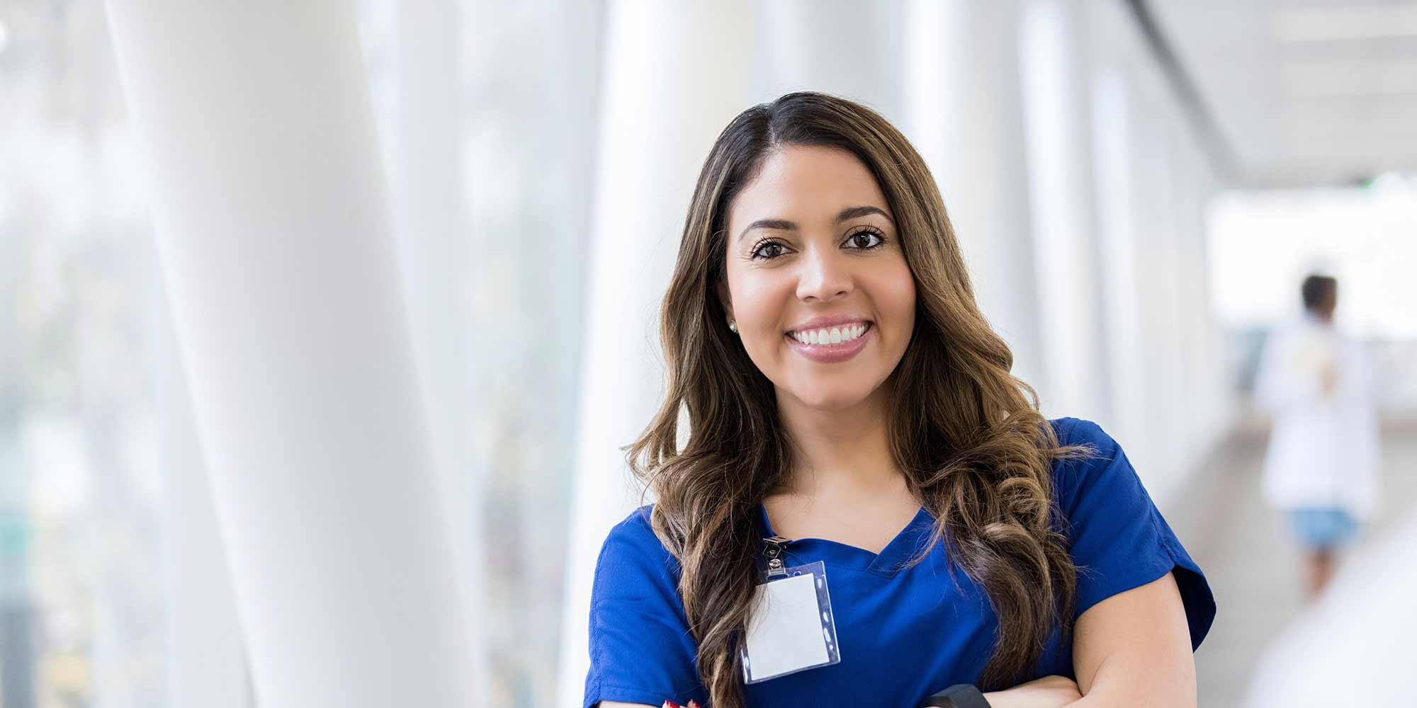 A woman stands in scrubs with her arms crossed in the middle of a hospital hallway