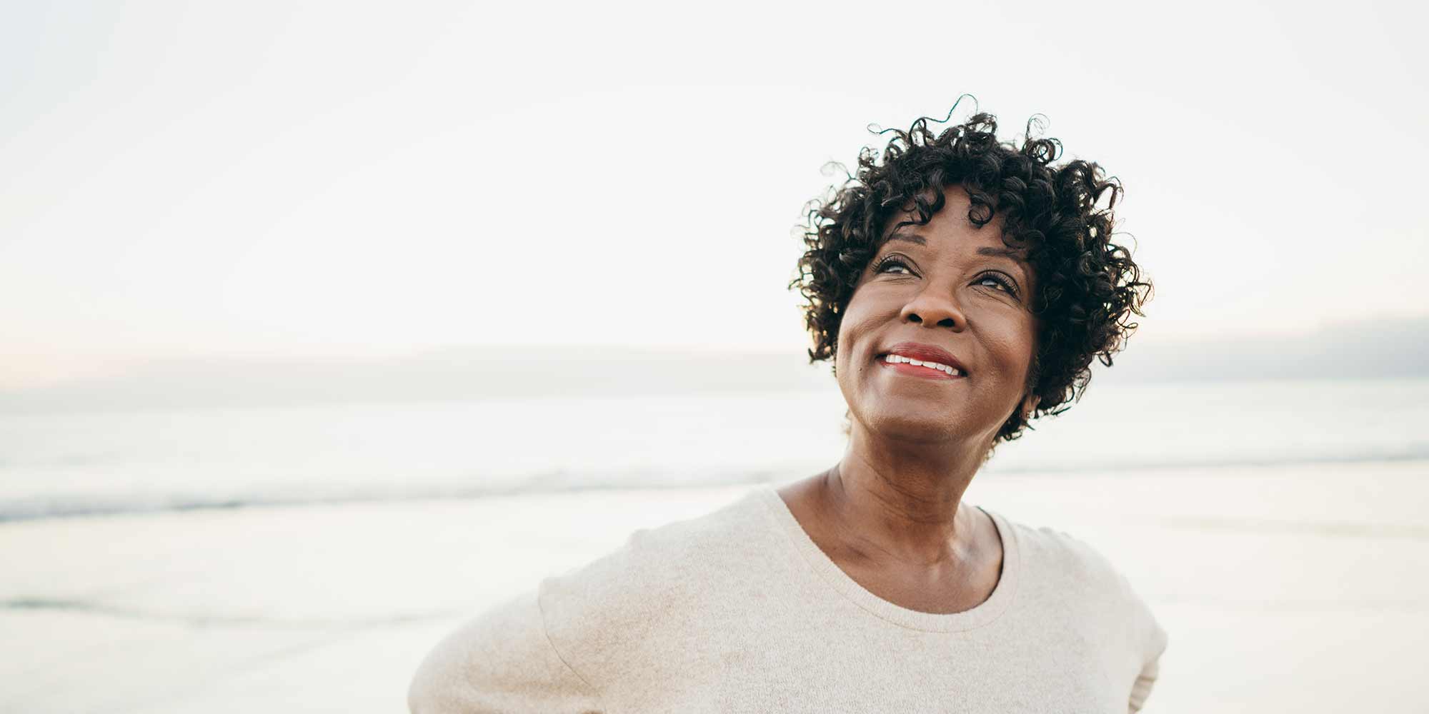 Middle-aged woman stands with the beach in the background, gazing and smiling slightly into the sky